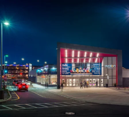 Family couple arriving at Avalon Mall entrance at night, warmly lit façade and busy car drop-off area