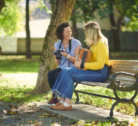 Two people enjoy a sunny day on a park bench, sharing smiles and holding a camera and an accordion.