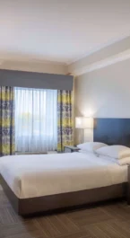 Bright hotel room with a double bed, desk, and natural light from a large window.