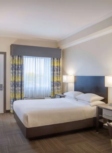 Bright hotel room with a double bed, desk, and natural light from a large window.