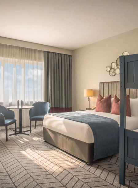 Family-friendly hotel room with double bed and blue bunk beds, seating area and warm natural light, visible table lamps and curtains