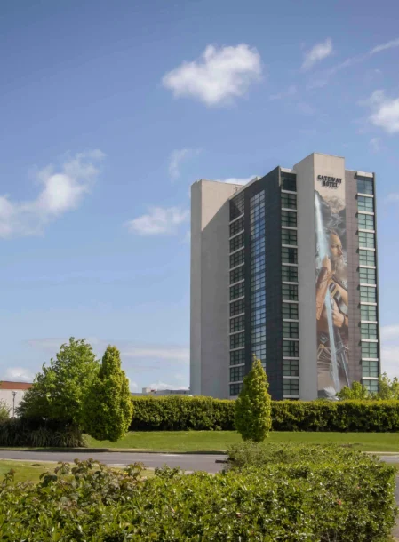 Modern tall hotel tower by a leafy roundabout, family enjoying bright sky and green grounds, sign reads "GATEWAY HOTEL" (88 characters)