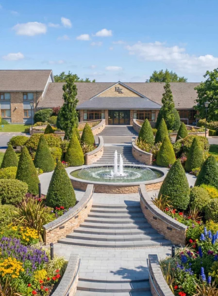 Family strolling past trimmed topiary and central fountain towards hotel entrance on sunny day