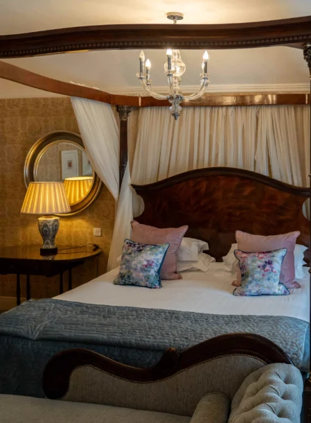 Romantic four-poster bedroom with family-friendly seating, floral cushions, lamps and chandelier (visible text: Allora CMS)